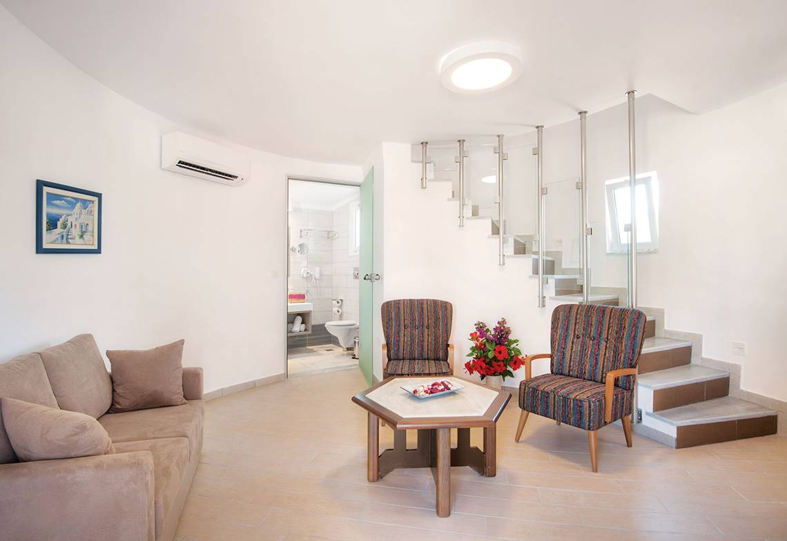Wohnzimmer des Eurovillage Achilleas Hotels mit Couch, Stühlen und Tisch neben einer Treppe.