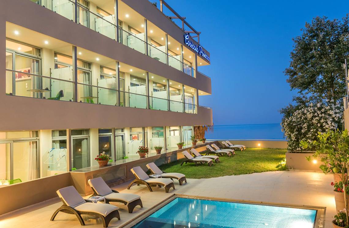 Außenansicht des Corissia Princess Hotels bei Abend, mit Swimmingpool und Sonnenliegen.