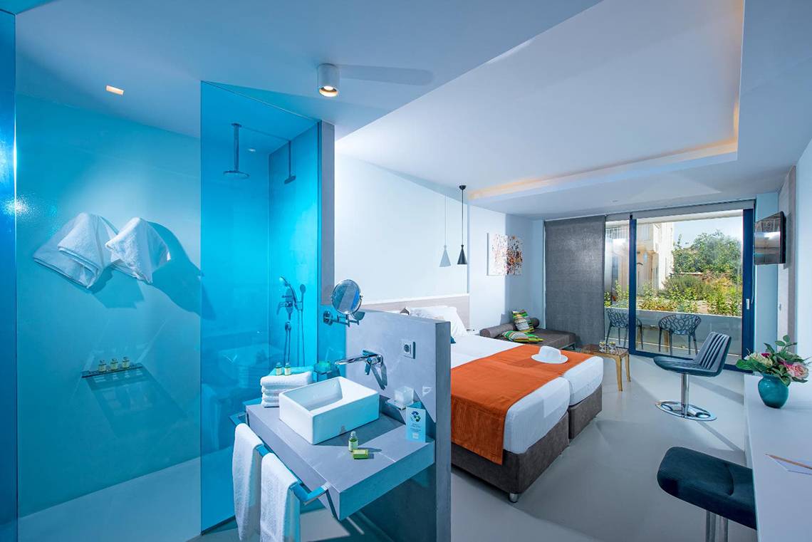Ein modernes Hotelzimmer im Corissia Princess Hotel mit blauen Wänden, einer Dusche, einem Waschbecken und einem Bett.