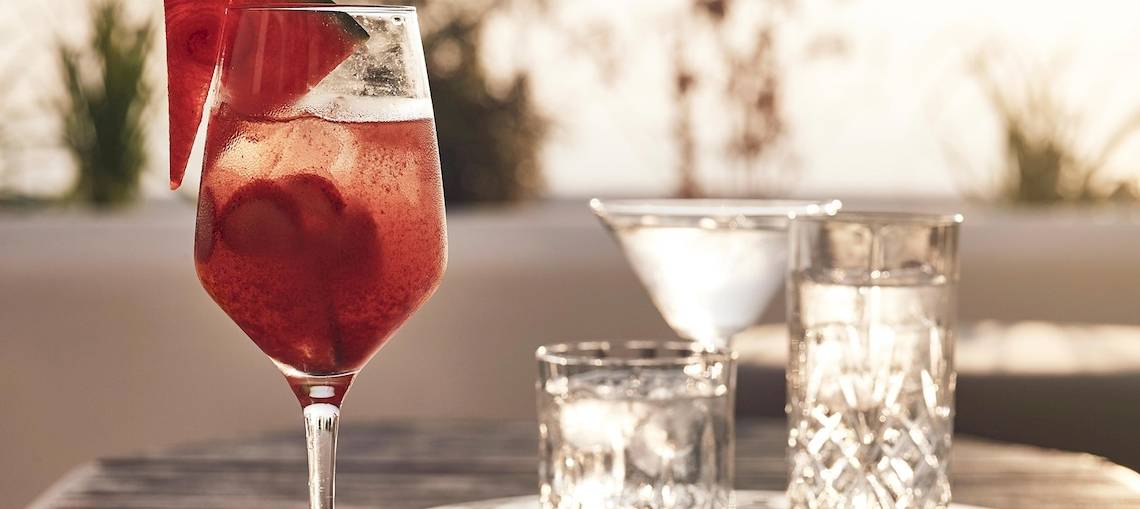 Ein Glas mit einem roten Cocktail und einem Stück Wassermelone auf einem Tisch mit weiteren Gläsern.