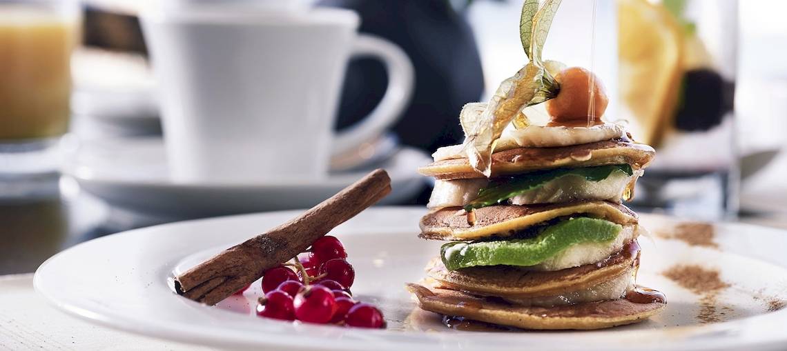 Frühstück im Santo Pure Oia Suites & Villas mit Pfannkuchen, Früchten und Zimtstange auf einem weißen Teller.