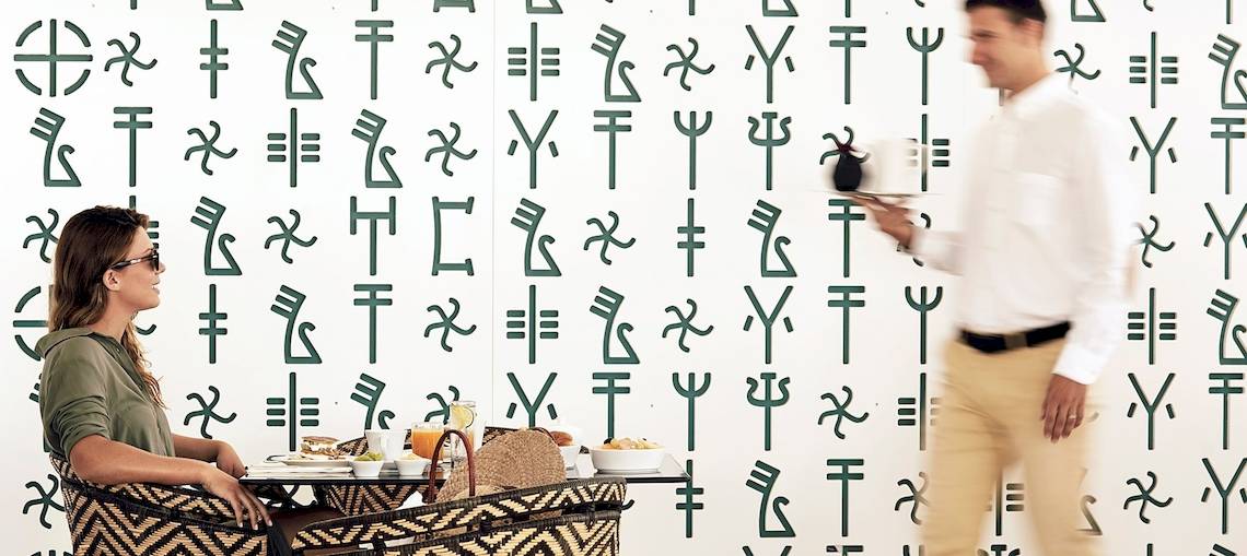 Eine Frau sitzt an einem Tisch in einem Hotel mit einer Wand voller grüner Symbole. Ein Mann geht vorbei.