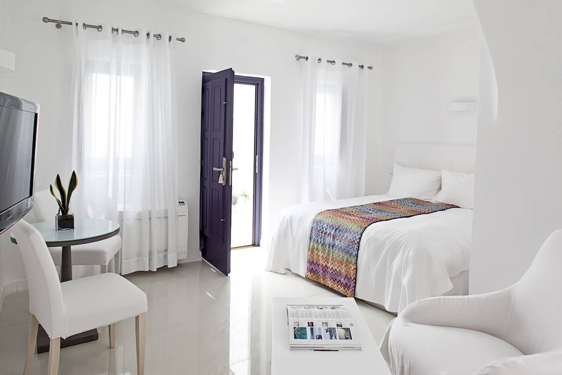 Innenansicht eines Zimmers im Katikies Chromata Santorini Hotel mit weißem Bett, Tisch und Fernseher