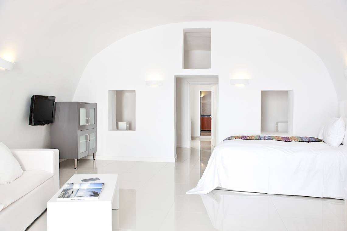 Innenansicht eines Schlafzimmers im Katikies Chromata Santorini Hotel mit weißem Bett, Couch, Tisch und Fernseher.