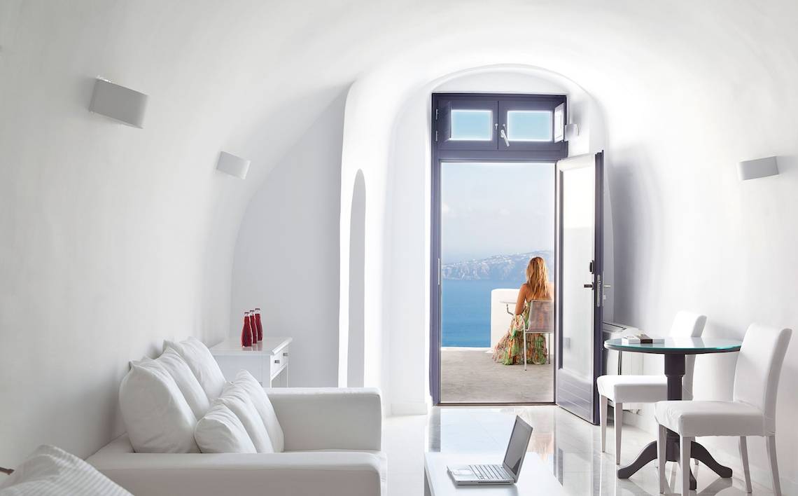 Ein helles Zimmer mit offenem Balkon und Meerblick, Möbel sind weiß und die Frau sitzt draußen.