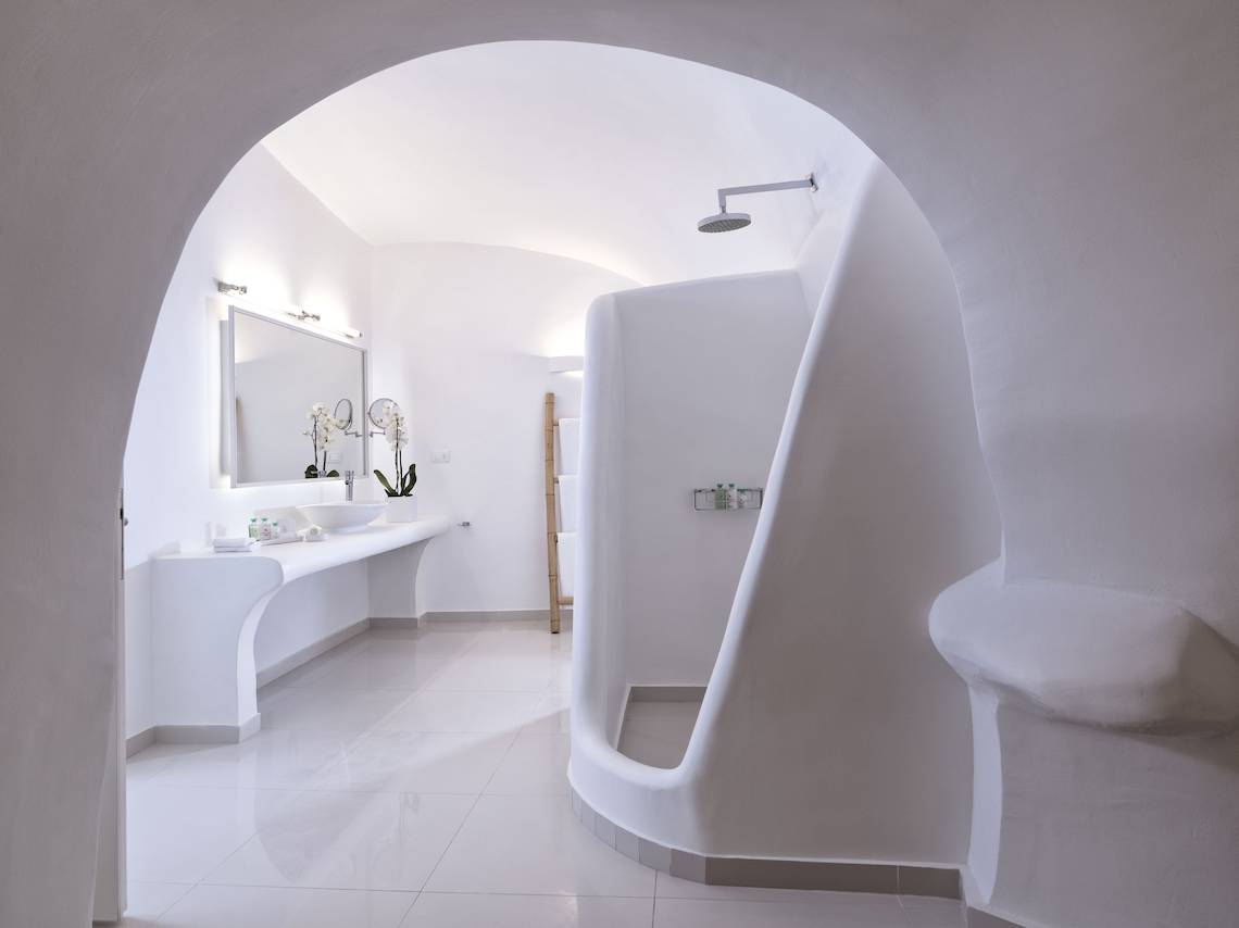 Badezimmer im Katikies Chromata Santorini Hotel mit weißer Architektur, Waschbecken und Dusche.