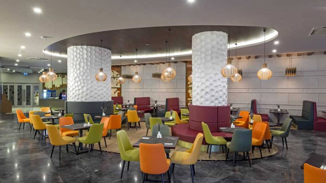 Innenansicht des Restaurants im Royal Seginus Hotel mit bunten Stühlen und Tischen, hohen Säulen und dekorativen Lampen.
