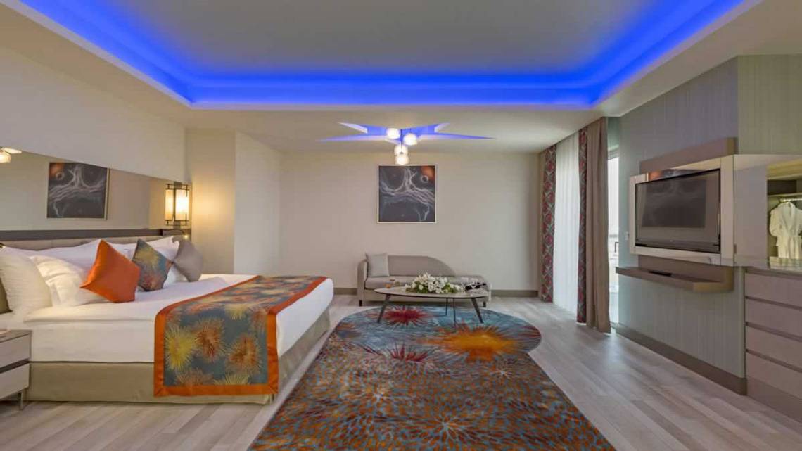 Innenansicht der Suite im Royal Seginus Hotel mit Bett, Wohnzimmerbereich und Fernseher.