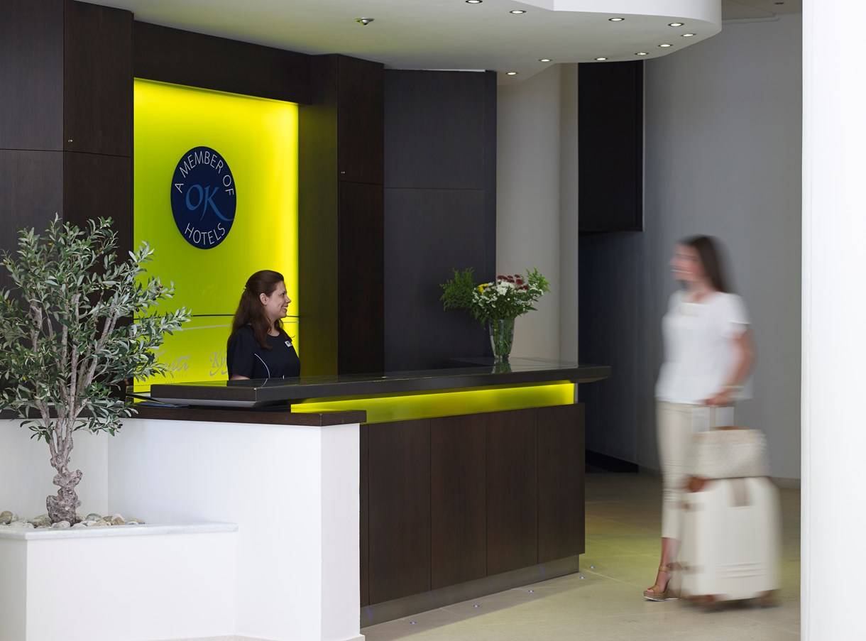 Empfang des Kriti Beach Hotels mit Rezeptionistin und Gast, die mit Gepäck unterwegs ist.