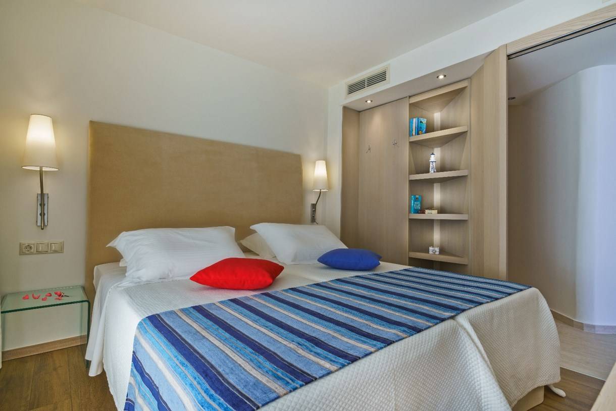 Bett im Kriti Beach Hotelzimmer mit zwei weißen Kissen, einem roten und einem blauen Kissen, und einer blau-weiß gestreiften Decke.