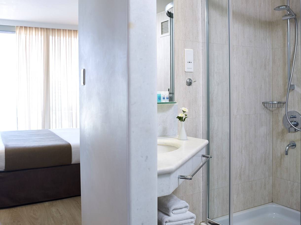 Badezimmer im Kriti Beach Hotel mit Dusche, Waschbecken und Bett im Hintergrund.
