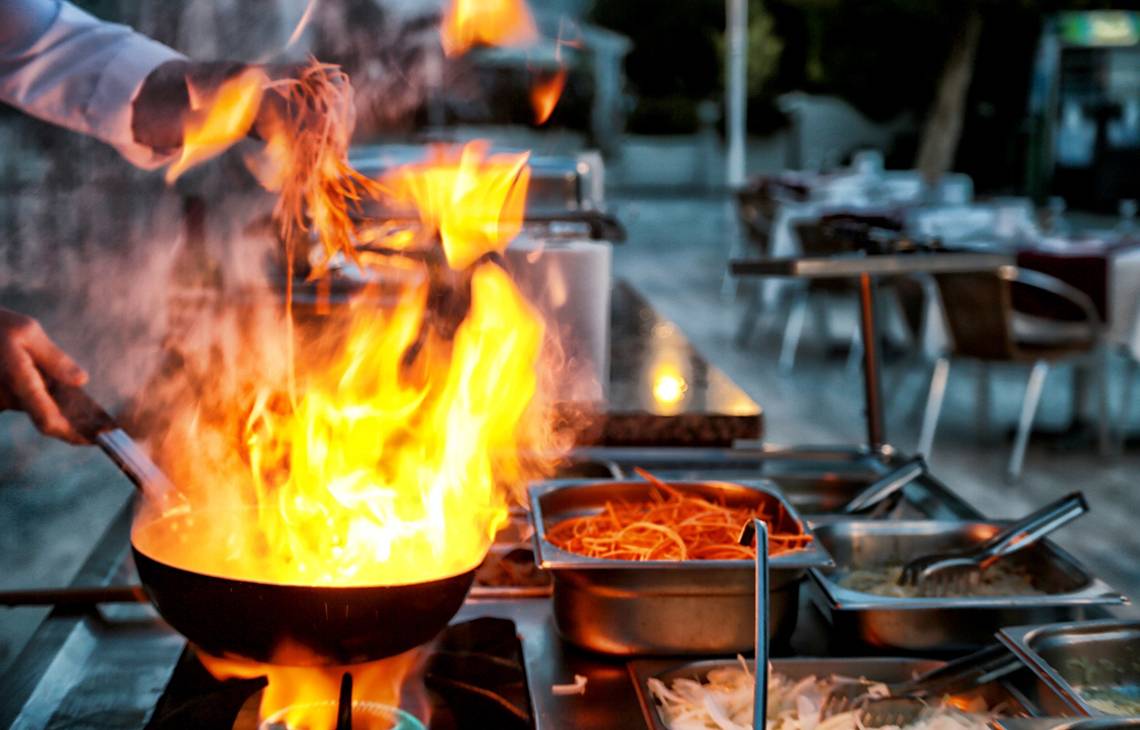 Ein Koch kocht mit Feuer in einer Pfanne auf einer Gasherd im Restaurant.