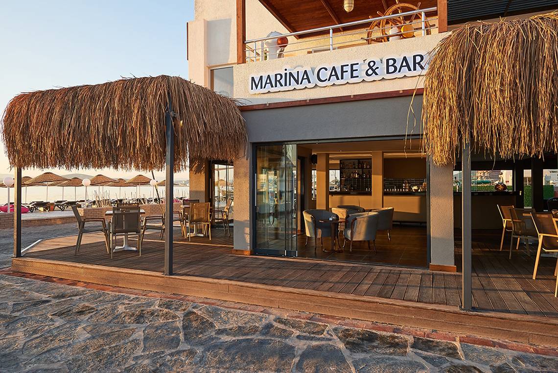 Außenansicht des Marina Cafe & Bar des Altin Yunus Hotels mit Strohdächern und Tischen im Freien.