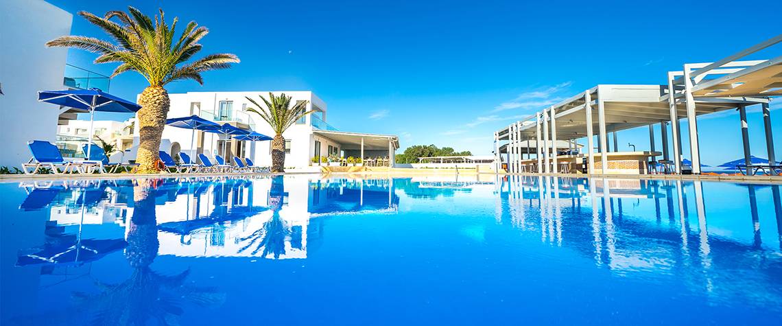 Außenpool des Adele Beach Hotels mit weißen Gebäuden, Sonnenliegen und Palmen im Hintergrund.
