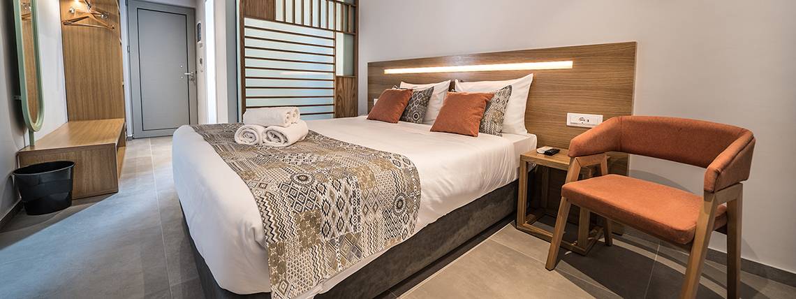 Schlafzimmer im Adele Beach Hotel mit Doppelbett, Dekokissen, Nachttisch, Stuhl und Eingangstür.