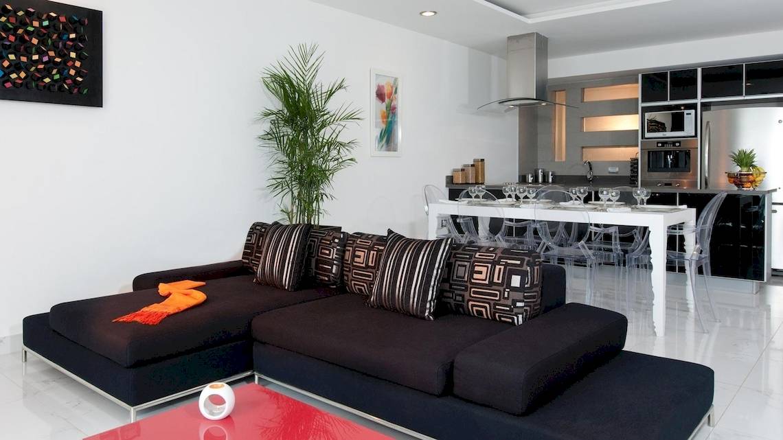 Modernes Wohnzimmer des Grand Sirenis Riviera Maya Resort mit schwarzem Sofa, weißem Esstisch und stilvoller Küche im Hintergrund.