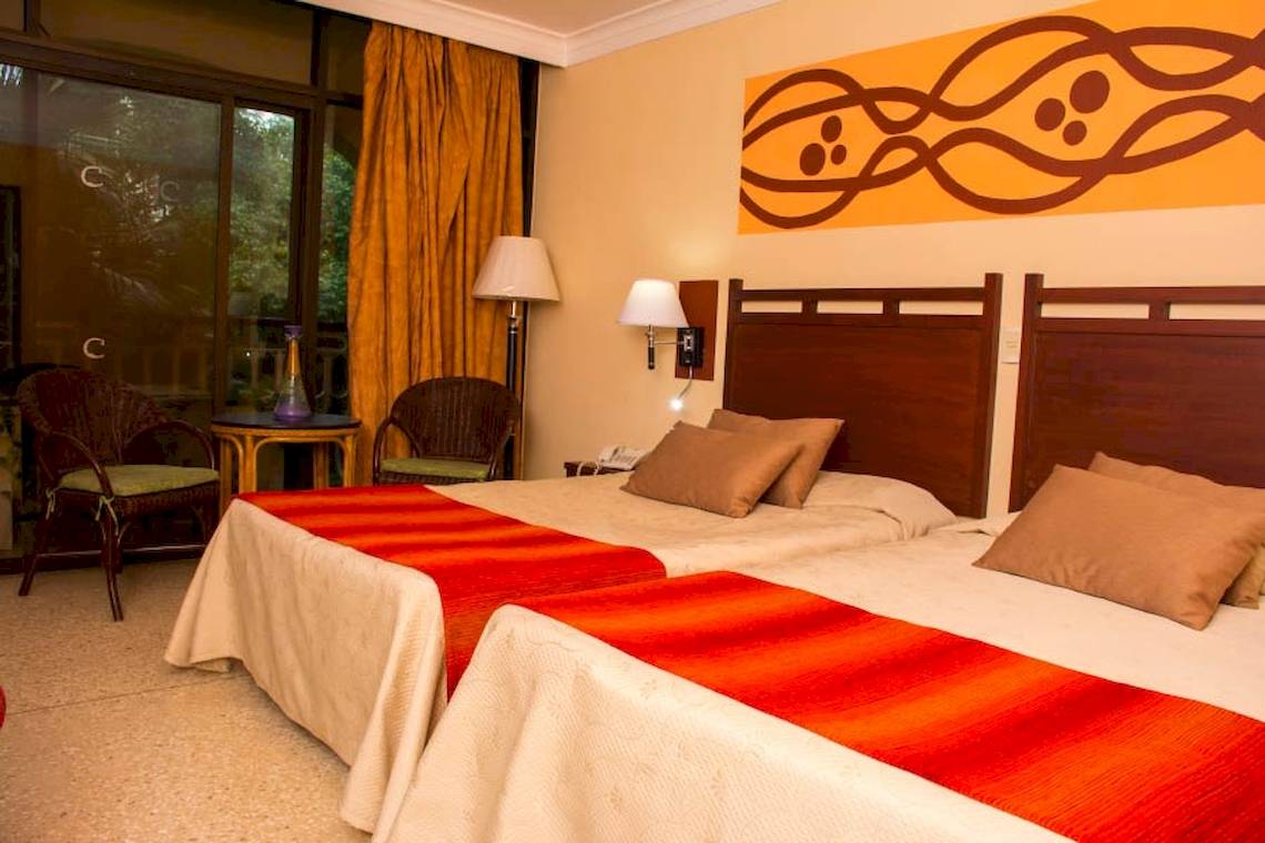Doppelbettzimmer im Roc Barlovento Hotel mit zwei Betten, Kopfteilen, Kissen, Decken, Tisch, Stühlen und Lampen.