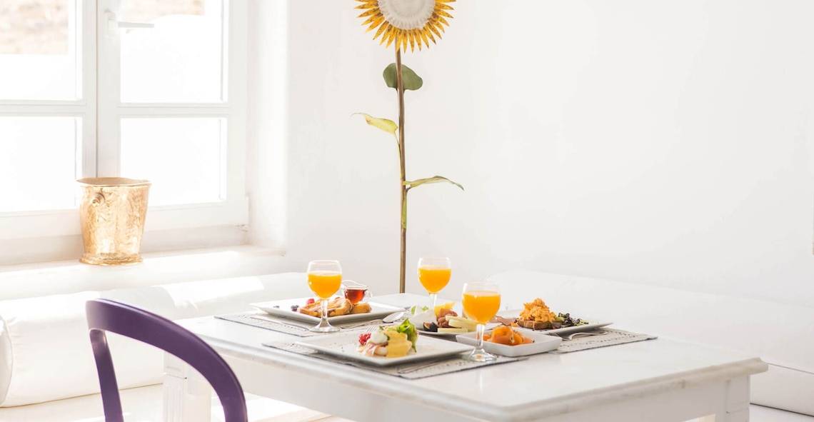 Frühstückstisch im Kivotos Mykonos Hotel mit Orangensaft, Speisen und einer Sonnenblume im Hintergrund.