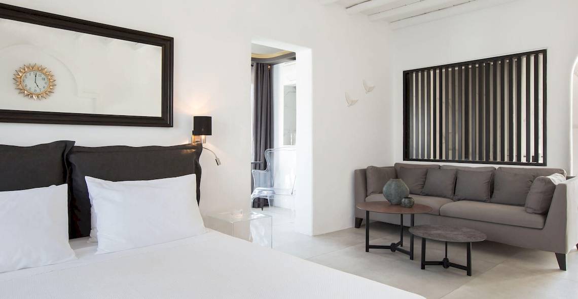 Ein modernes Hotelzimmer von Kivotos Mykonos mit weißem Bett, grauem Sofa, Tisch und Uhrspiegel.