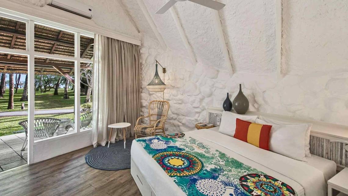 Innenansicht eines Zimmers im La Pirogue Mauritius Hotel mit weißem Bett, Holzboden und Glastür.