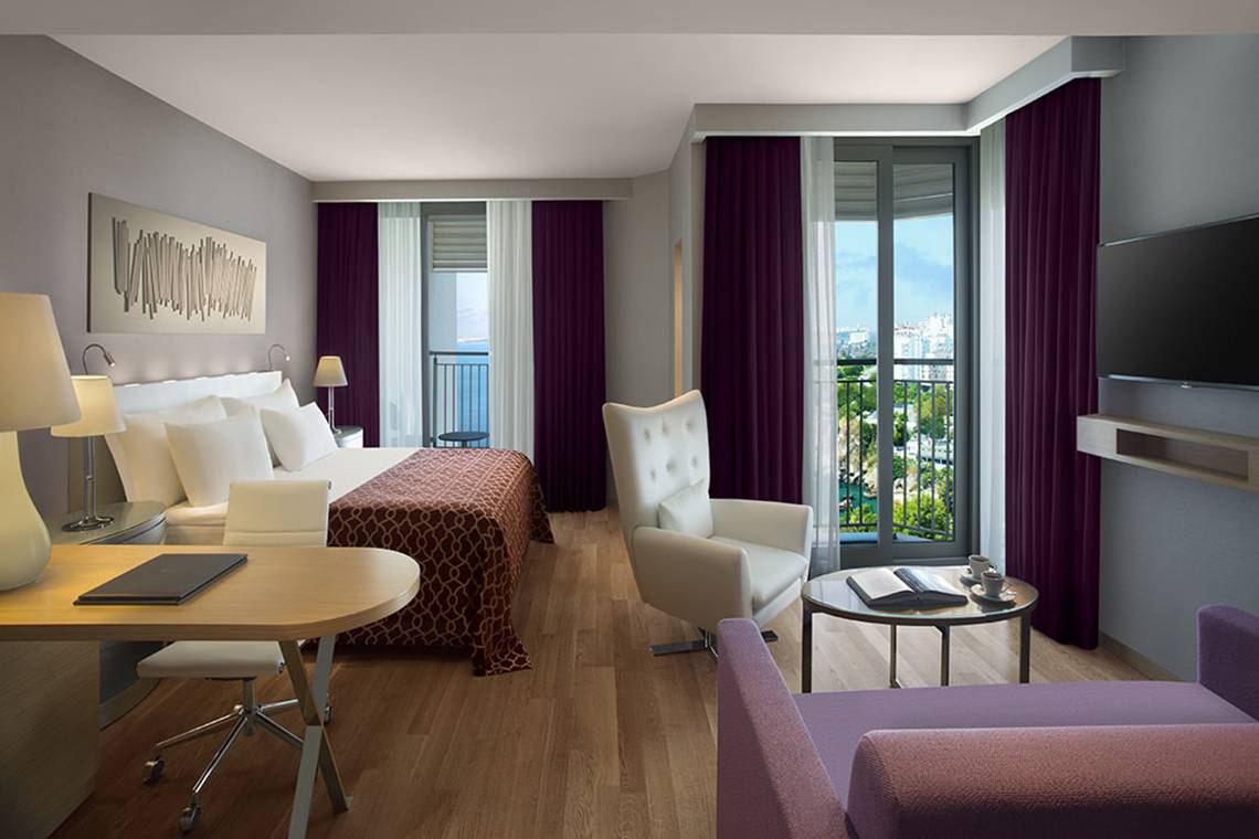 Innenansicht eines Zimmers im Akra Hotel mit Bett, Schreibtisch und Balkon.