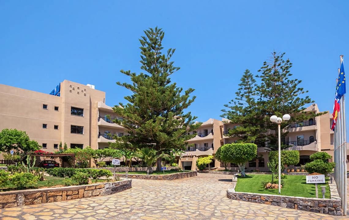 Außenansicht des Themis Beach Hotels mit einem gepflasterten Eingangsbereich, hohen Bäumen und gepflegten Gärten.