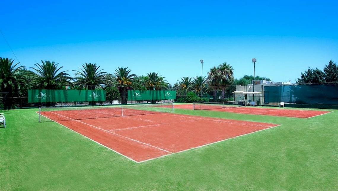 Tennisplätze im Freien im Aquila Rithymna Beach Hotel, umgeben von grünem Rasen und hohen Bäumen.