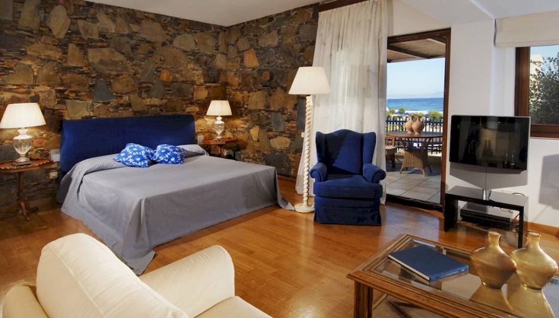 Schlafzimmer im Aquila Rithymna Beach Hotel mit Steinwänden, einem Bett, zwei Sesseln und einem Fernseher.