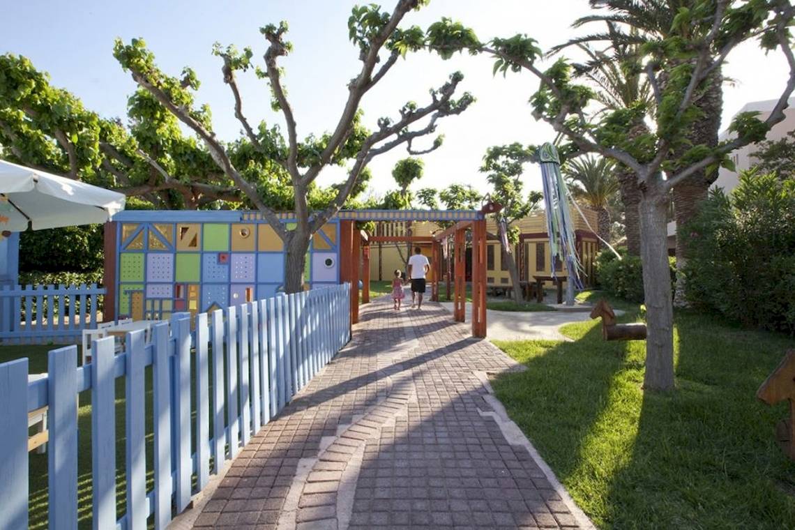 Kinderspielplatz im Freien am Aquila Rithymna Beach Hotel mit bunten Gebäuden und Bäumen.