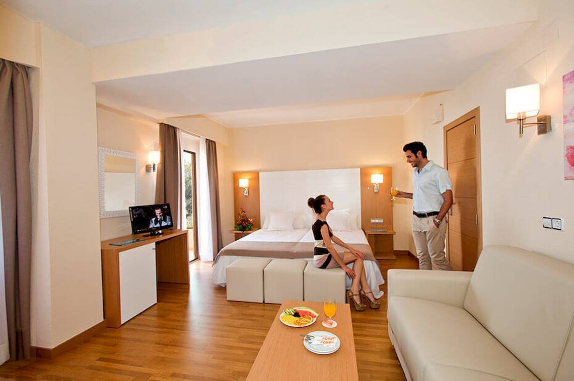 Innenansicht der Suite im HSM Venus Playa Hotel mit einer Frau auf einem Hocker und einem Mann, der ein Getränk serviert.