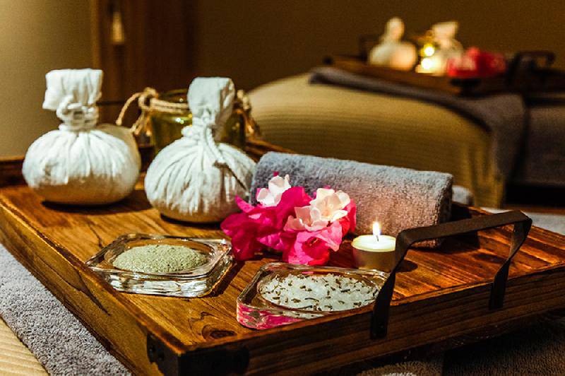 Ein Holztablett mit Spa-Zubehör im Crystal Springs Beach Hotel, einschließlich zweier weißer Beutel, einem Handtuch, Blumen, Kerzen und kleinen Glasbehältern.