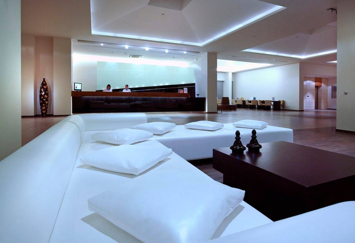 Hotellobby mit weißen Sofas, Kissen und einem dunklen Tisch vor einer Rezeption.