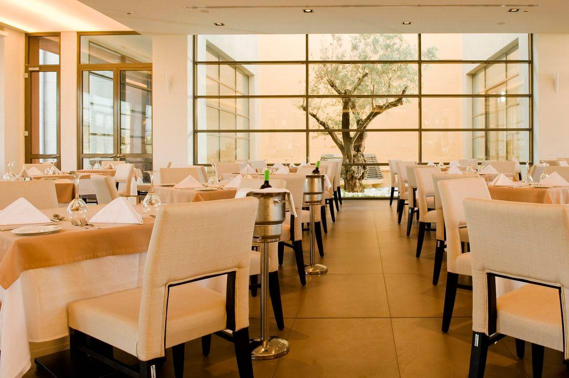 Innenansicht eines Restaurants im Astir Odysseus Kos Resort & Spa mit gedeckten Tischen und Blick auf einen Olivenbaum.