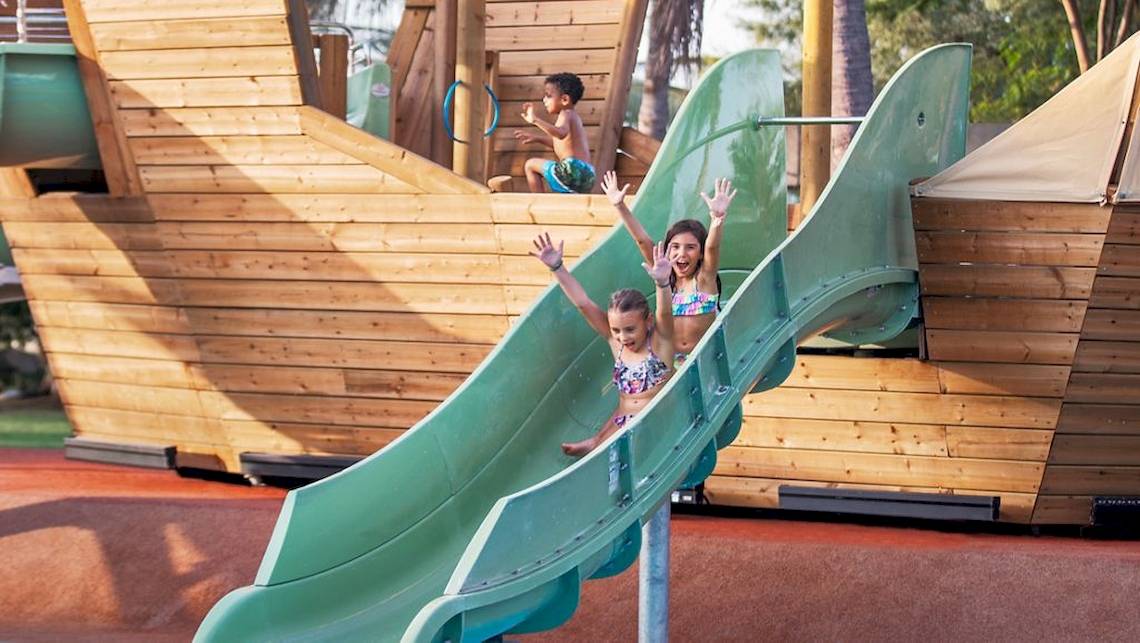 Kinder rutschen im Freizeitpark, zwei Mädchen und ein Junge im Hintergrund.