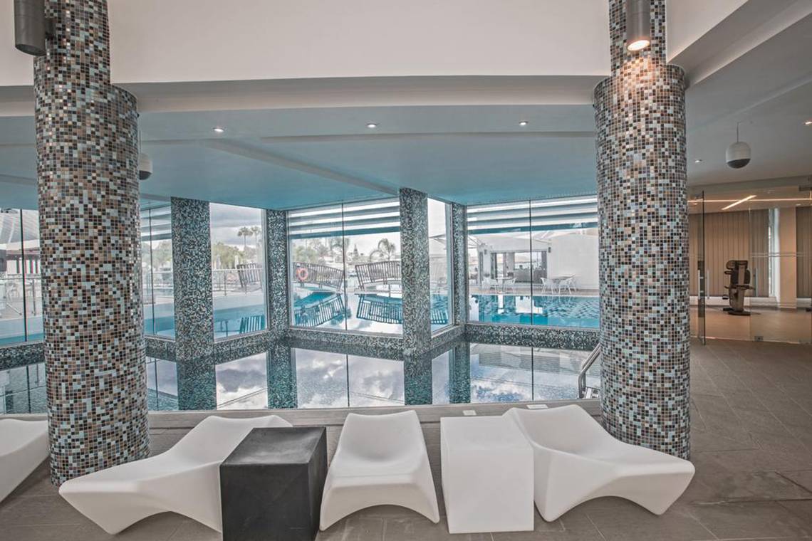 Innenpool-Ansicht im Vassos Nissi Plage Hotel mit weißen Sesseln und gefliesten Säulen.