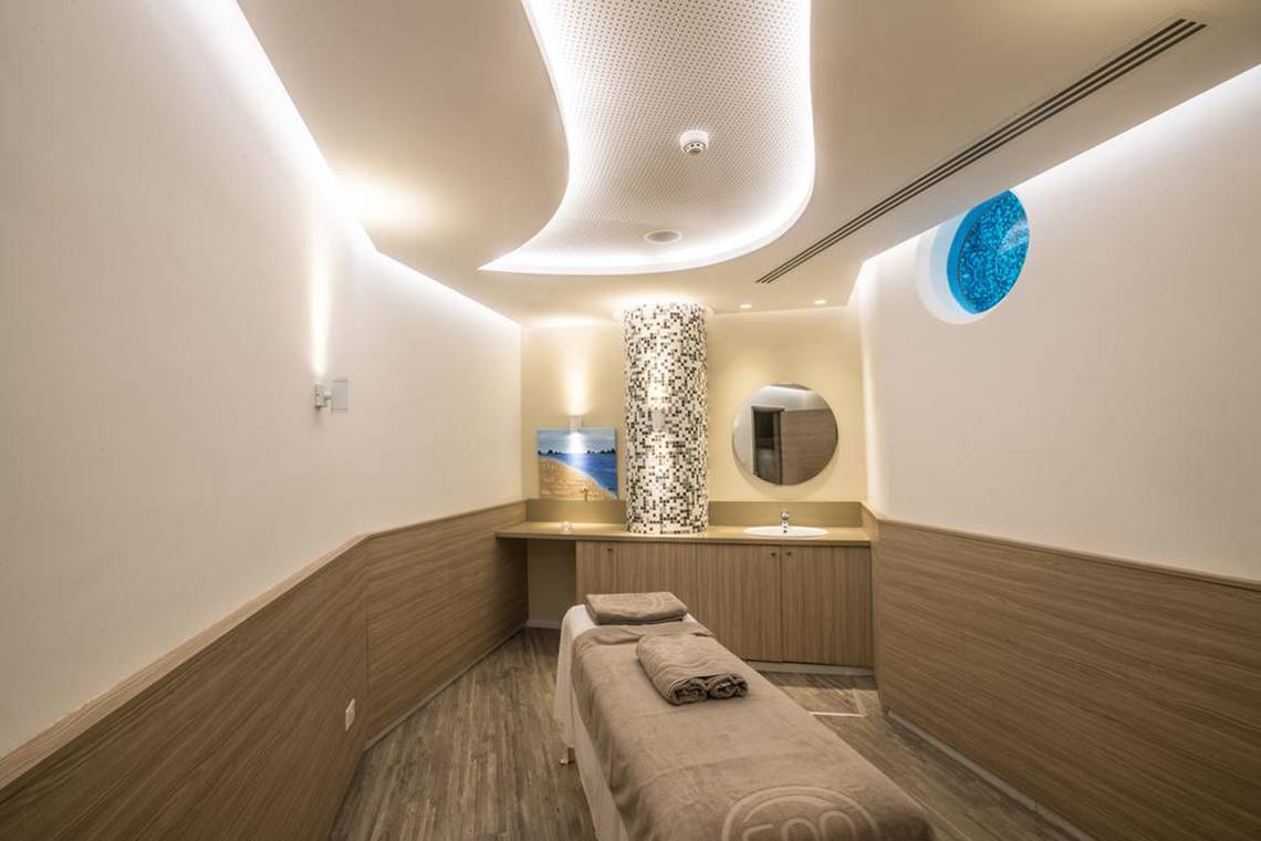 Innenansicht des Massageraums im Vassos Nissi Plage Hotel mit einem Behandlungstisch, einem Spiegel und einer blauen Glasscheibe.