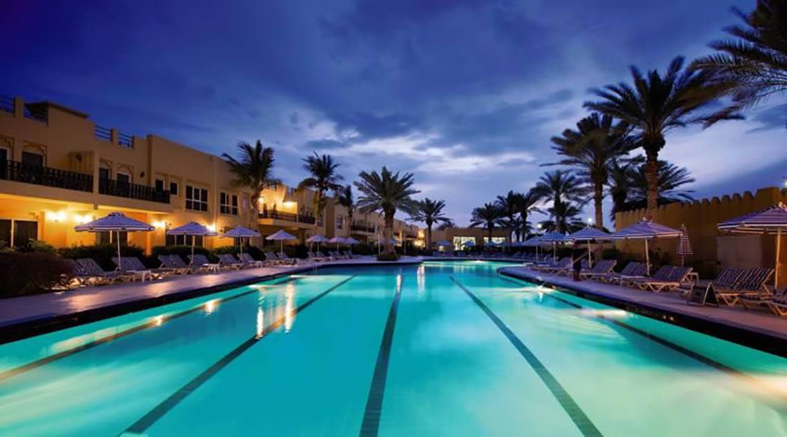 Al Hamra Village Hotel Pool bei Nacht mit Lounge-Stühlen und Sonnenschirmen.