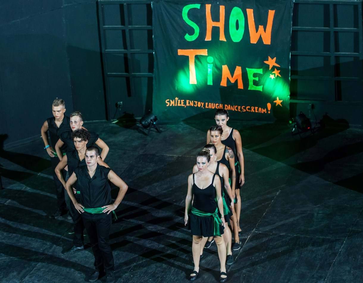 Tanzgruppe auf der Bühne unter einem 'Show Time'-Banner, das mit grünem Licht beleuchtet ist.