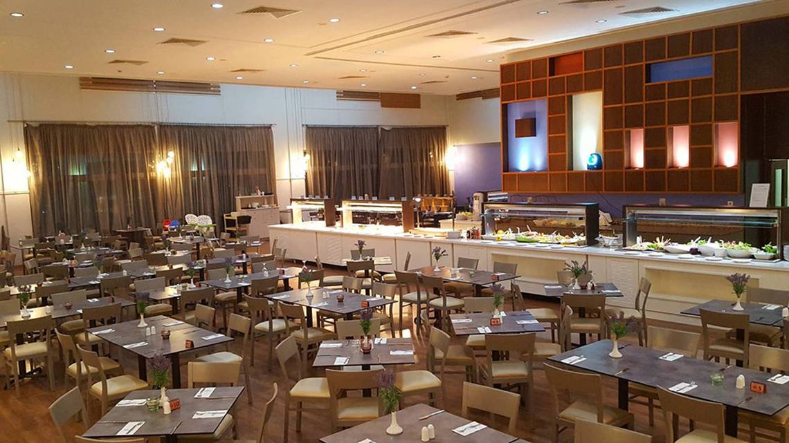 Innenansicht des Restaurants im Aktea Beach Village Hotel mit vielen gedeckten Tischen und einer langen Theke im Hintergrund.
