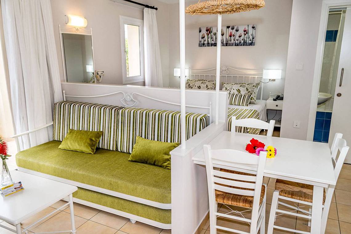 Aktea Beach Village Zimmer mit grünem Sofa, Tisch und Stühle, weißer Wand und Fenster.