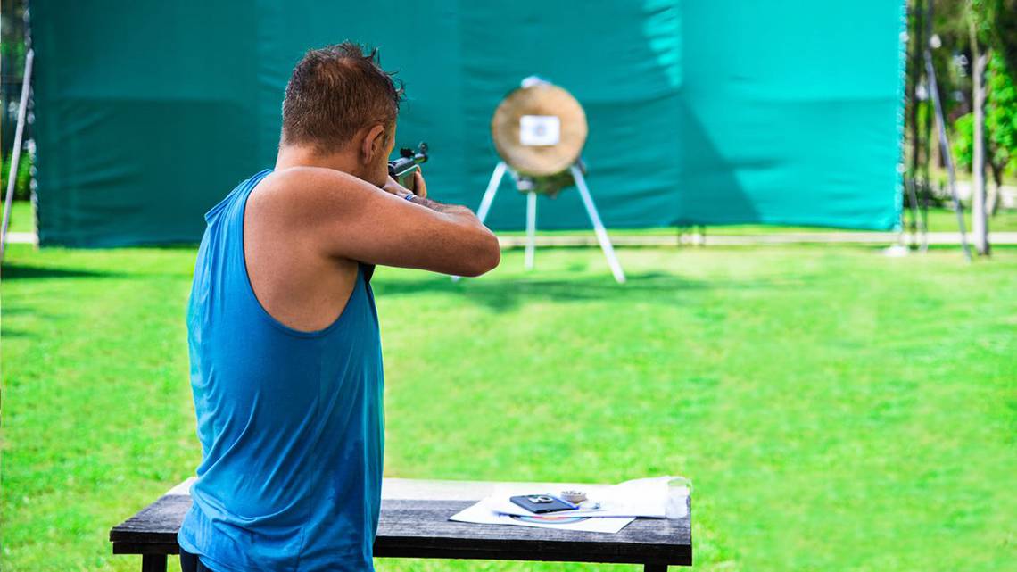 Ein Mann in einem blauen Tanktop zielt mit einer Waffe auf eine Zielscheibe auf einer Wiese.