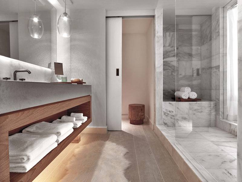 Innenansicht des Badezimmers im 1 Hotel South Beach mit Marmorfliesen, Glasdusche und Holzmöbeln.