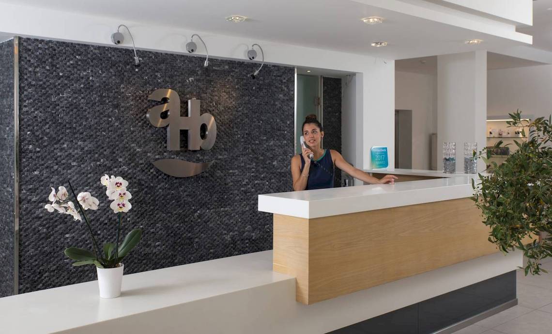 Rezeptionistin des Acharavi Beach Hotel telefoniert, Empfangstresen im Vordergrund, Logo an der Wand im Hintergrund.