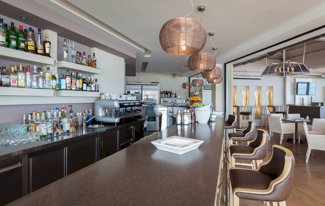 Innenansicht der Bar im Acharavi Beach Hotel mit langen Theke, Stühlen und Flaschen.
