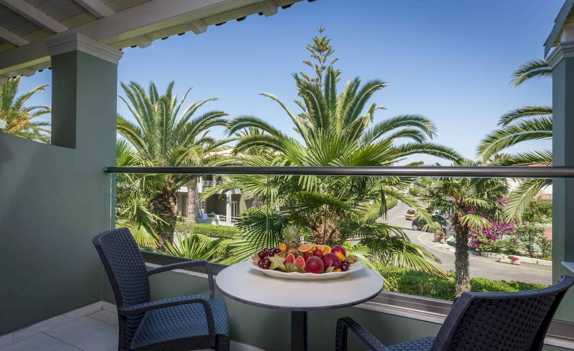 Balkon des Acharavi Beach Hotel mit Obstplatte und Aussicht auf Palmen und Straße.