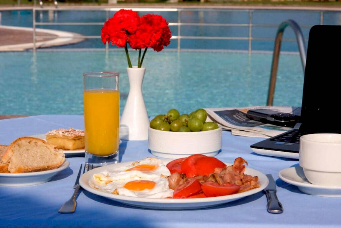 Frühstück am Pool des Plaza Beach Hotels mit rotem Blumenstrauß, Orangensaft, Spiegeleiern, Tomaten, Speck und Laptop.