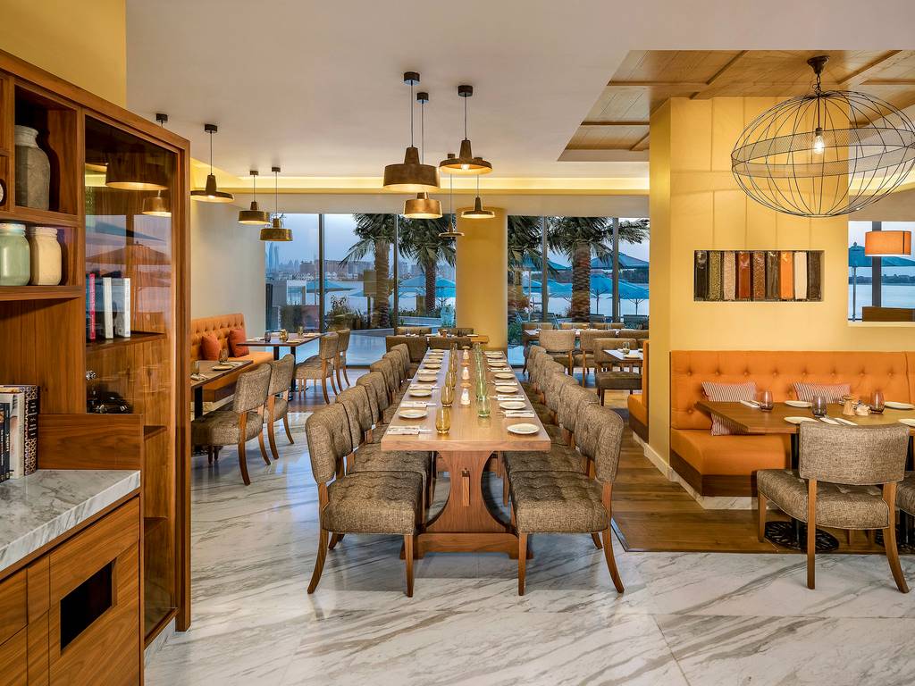 Innenansicht des Restaurants im Retreat Palm Dubai MGallery by Sofitel mit langer Tafel und Aussicht auf Palmen und Meer.