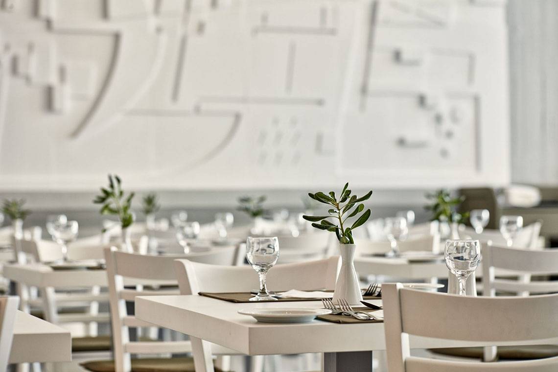 Restaurant-Interieur des Sentido Unique Blue Resort & Villas mit weißen Tischen und Stühlen, gedeckten Tischen und Pflanzen in Vasen.