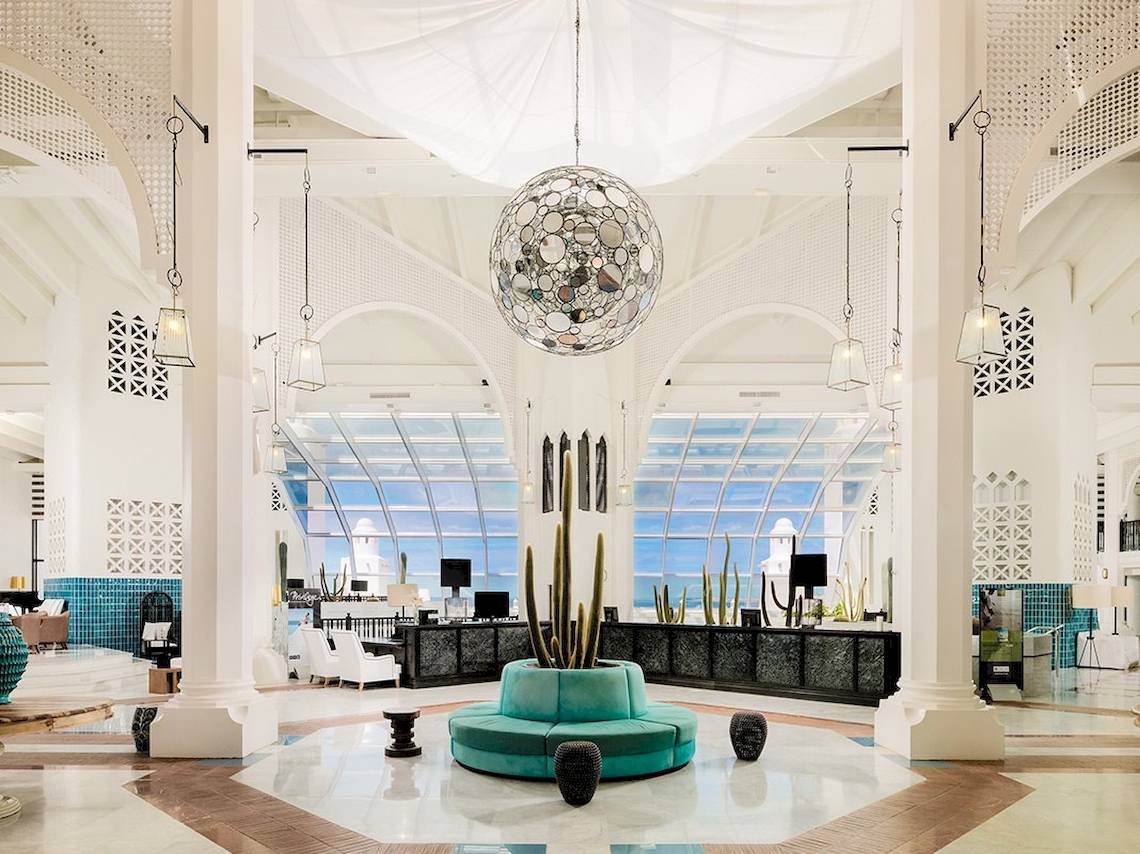Innenansicht der Lobby des H10 Timanfaya Palace mit zentralem Kaktus in blauem Topf und Spiegel-Pendelleuchte.