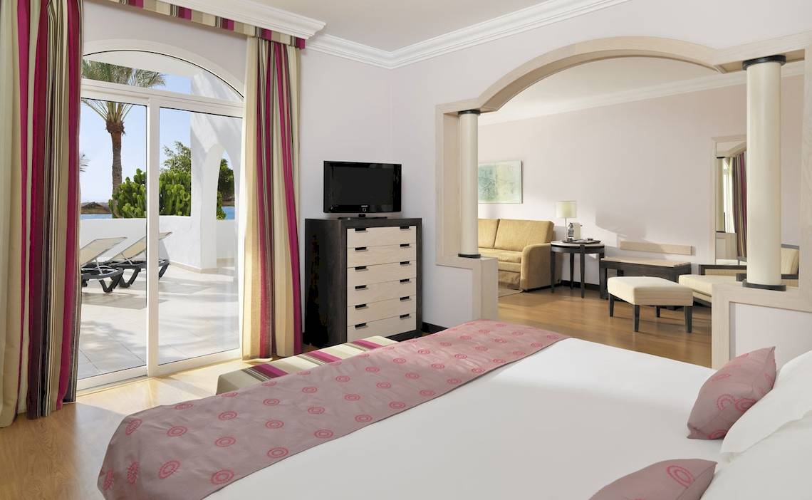 Innenansicht der Suite im H10 Timanfaya Palace Hotel mit Bett, Fernseher, und Blick auf die Terrasse.